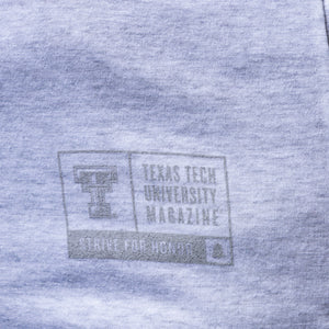 Close-up of a heather gray fabric surface with a faded gray printed Texas Tech University Magazine logo and text.