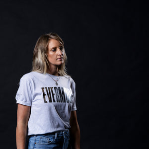 A woman with blonde hair wearing a light gray graphic t-shirt with the word 'EVERMIORE' printed in black across the chest, paired with blue jeans, set against a solid black background.
