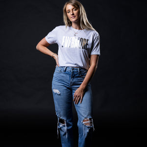 A woman modeling a light gray graphic t-shirt with 'EVERMORE' printed in metallic silver across the chest, paired with distressed blue jeans, posed against a solid black background.