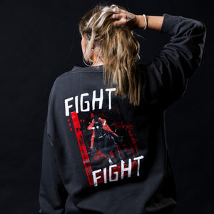 A woman viewed from behind wearing a dark charcoal gray sweatshirt with a large graphic print on the back. The graphic features the words 'FIGHT RAIDERS FIGHT' and an archival image of the Masked Rider