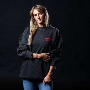 A woman modeling a black crewneck sweatshirt with a red 'TECH'  pennant logo on the chest, paired with blue jeans, set against a solid black studio background.