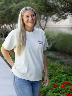 A smiling blonde woman wearing a white short-sleeve t-shirt with 'TEXAS TECH' printed in red on the left chest, paired with blue jeans, standing outdoors on a paved path next to green foliage and red flowers.