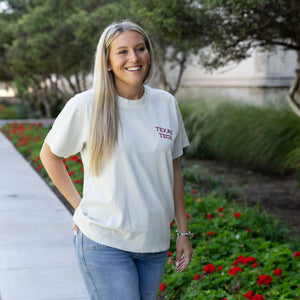 A smiling blonde woman wearing a white short-sleeve t-shirt with 'TEXAS TECH' printed in red on the left chest, paired with blue jeans, standing outdoors on a paved path next to green foliage and red flowers.