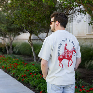A man wearing a white t-shirt with a red graphic of a cowboy on a horse and the text 'NOTIN' PLAIN ABOUT IT' on the back, standing outdoors on Texas Tech Campus